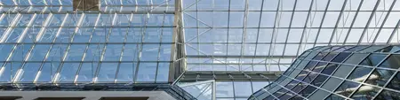 Doorkijk naar een modern gebouw met een groot glazen dak en gevelconstructie. De structuur bestaat uit metalen spanten en transparante panelen, met rechts een gebogen glazen vorm in een rasterpatroon.