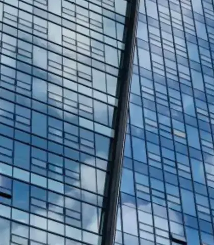 Hoekaanzicht van een moderne glazen wolkenkrabber tegen een deels bewolkte blauwe lucht. De gevel weerspiegelt de wolken, wat een patroon van licht en lucht op het gebouw creëert.